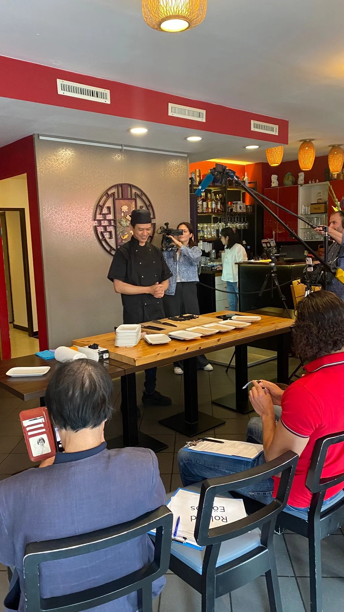 Bühne im China Restaurant Yung — Chi Kei Yung in schwarzer Kochjacke vor leeren Tellern, Kamerateam und sitzende Zeugen mit Klemmbrett