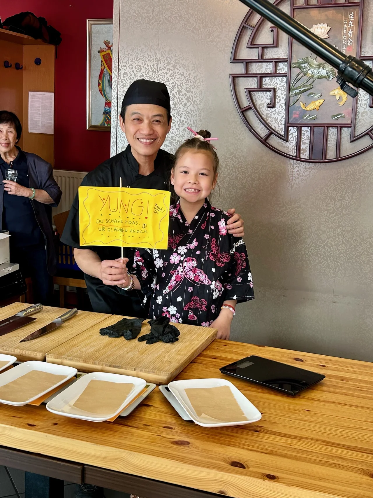 Chi Kei Yung im Restaurant kurz vor dem Versuch, daneben ein Mädchen mit selbstgemaltem gelben Schild ‚YUNG! Du schaffst das'