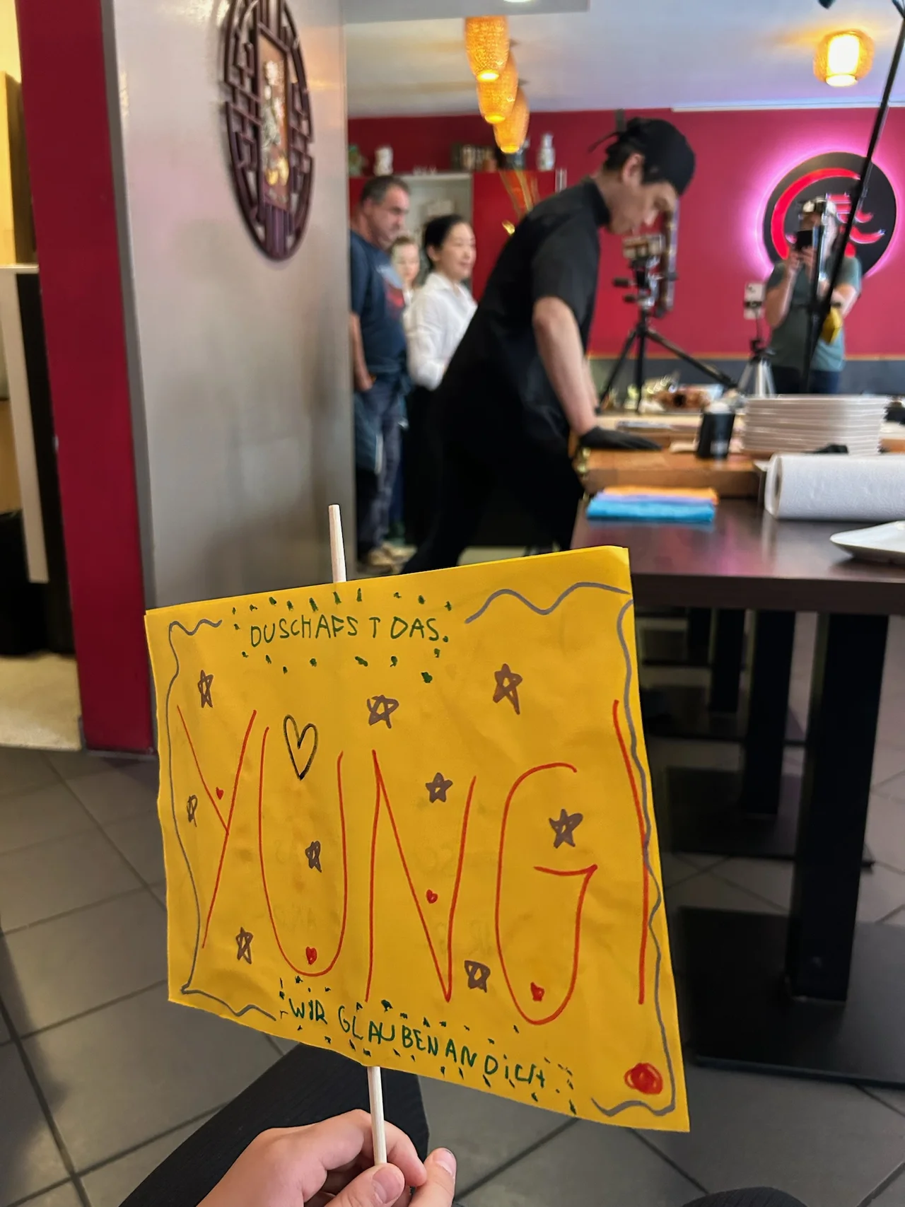 Selbstgemaltes gelbes Kindermal-Schild ‚YUNG! Wir glauben an dich' im Vordergrund, Chi Kei Yung im Hintergrund tranchierend, Kamerateam dokumentiert den Versuch