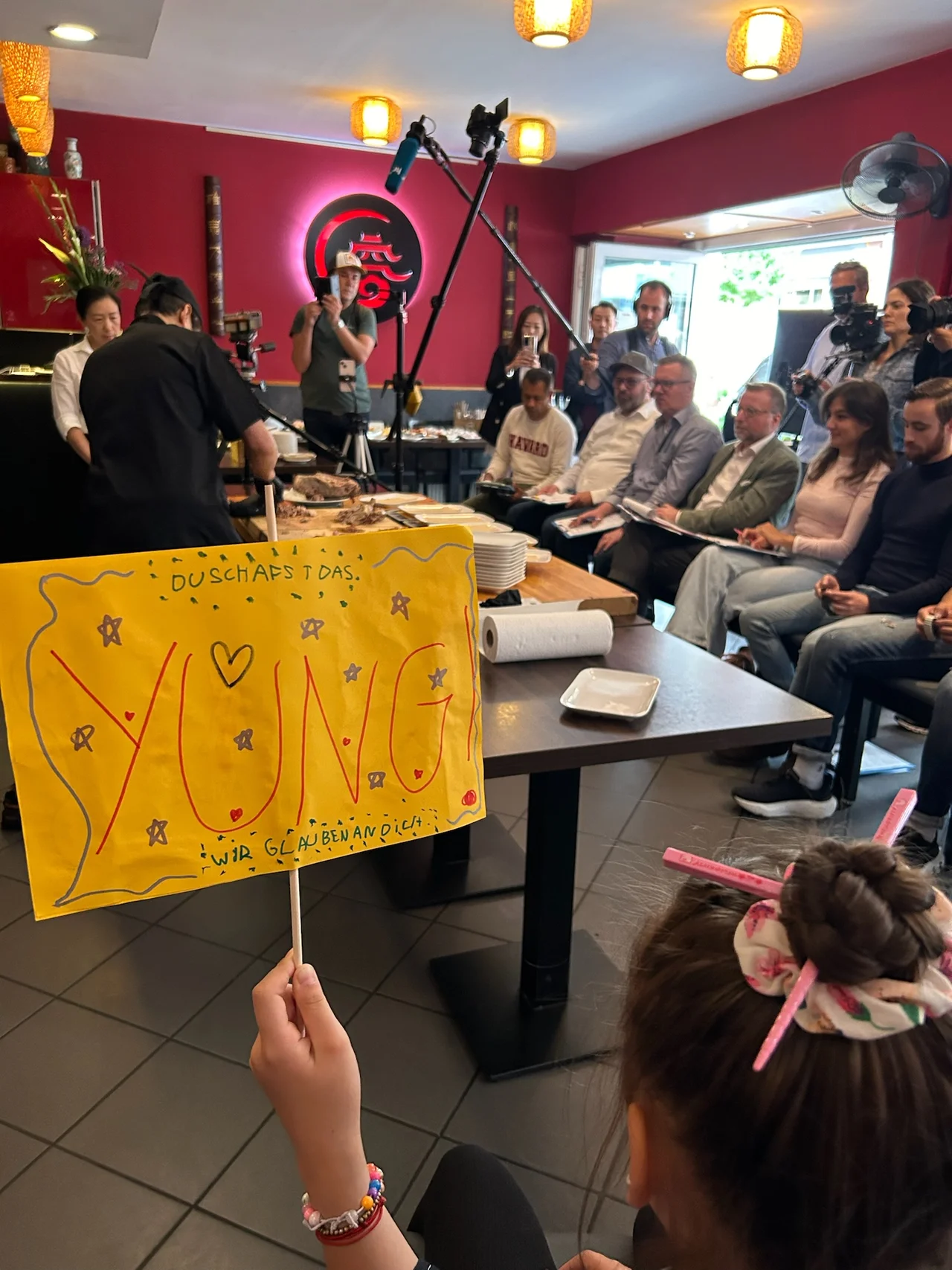 Voller Saal mit Pressekorps und Gästen — gelbes Kinderschild im Vordergrund, Chi Kei Yung im Hintergrund am Bühnentisch