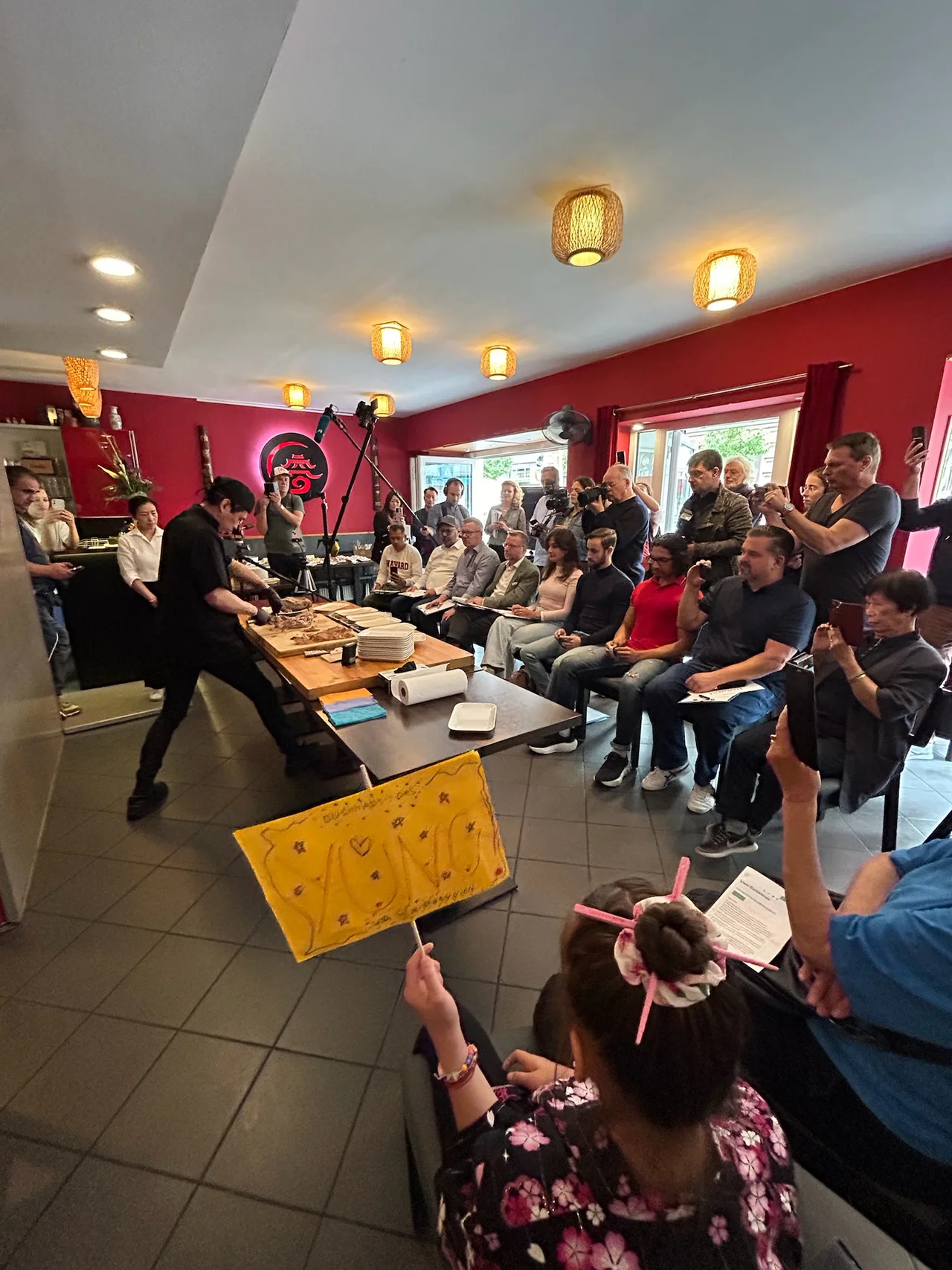 Chi Kei Yung wiegt Portionen, der Saal applaudiert, Mädchen mit Schild im Vordergrund